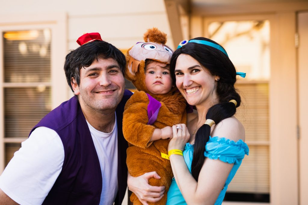 Three guests dressed as Aladdin, Abu, and Jasmine from Aladdin. The guest on the right is dressed as Aladdin with a small red hat, purple vest, and white shirt. The guest in the middle (being held by the guest dressed as Jasmine) is wearing an Abu onesie with a purple vest. The guest dressed as Jasmine has a jeweled teal headband, long black hair, and teal shirt.