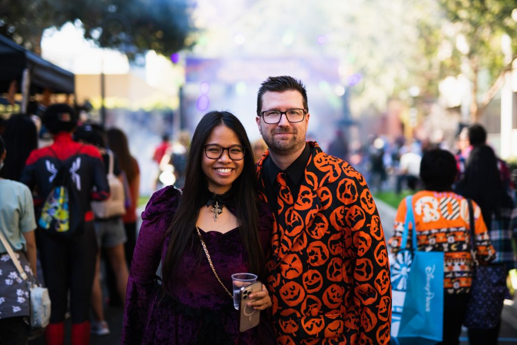 Two guests smiling in front of the Studio Lot activities. The guest on the left has long black hair, and is wearing glasses and a purple dress and black cape. The guest on the right has short brown hair, and is wearing glasses and a black suit decorated with bright orange pumpkins.