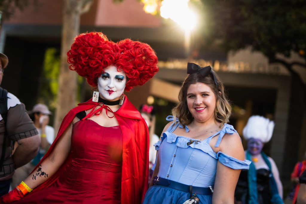 Two guests dressed as The Queen of Hearts and Alice from the Tim Burton’s Alice in Wonderland. The guest on the left has bright red hair made of roses, white face paint, bright blue eye shadow, and red lips drawn in a heart. The guest’s earrings are playing cards and they’re wearing a bright red dress and cape. The guest on the right has a black bow tied in their blond hair, and is wearing a light blue top with a dark blue skirt.