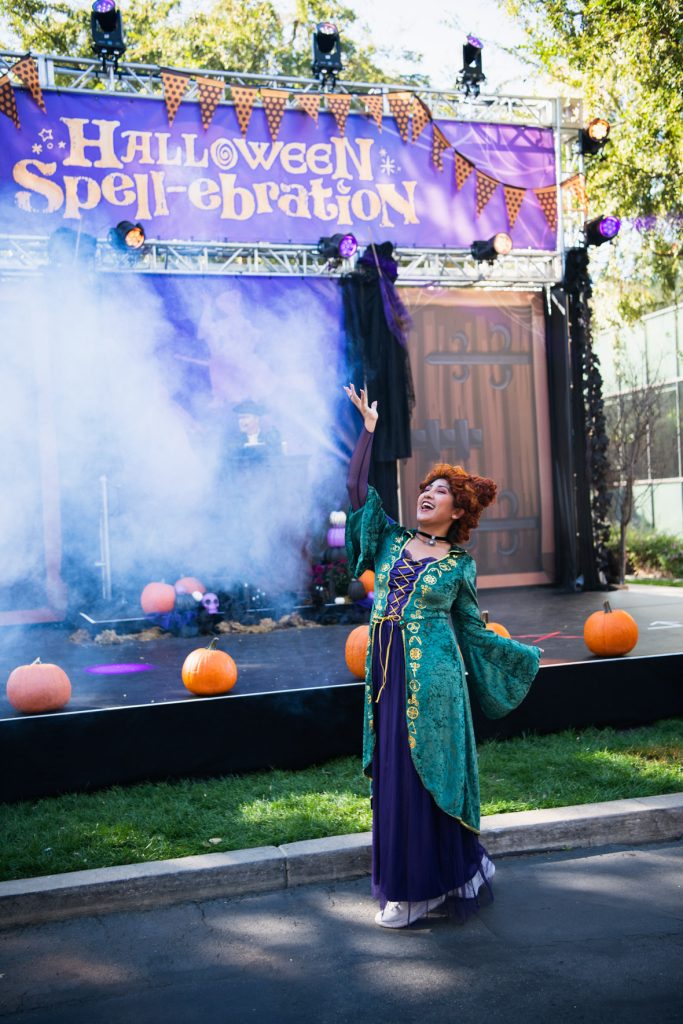 A guest dressed as Winifred Sanderson from Hocus Pocus posing in front of DJ Clark’s stage. The guest has a purple dress, green overcoat with gold designs, and red curly hair. The stage behind has pumpkins lining it with a banner at the top with the Halloween Spell-ebration logo in purple and yellow. The stage is covered in fog.