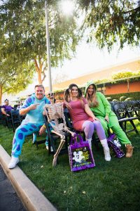 Three guests sitting with a skeleton holding a trick-or-treat back. The guest on the left is pointing to the skeleton and is dressed in a Sully onesie from Monsters, Inc. The guest sitting to the right of the skeleton is dressed as Boo with pig tails, a pink dress, and purple leggings. The guest sitting to the right is wearing a Mike Wazowski onesie. The trick-or-treat back the skeleton is holding is purple with a Mickey Mouse ghost and “Happy Halloween” written in green.