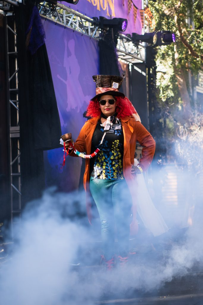 A guest dressed as The Mad Hatter from Tim Burton’s Alice In Wonderland emerging from the fog, walking across the Halloween Spell-ebration stage. The guest has a brown top hat with a pink ribbon, red curly hair and black sunglasses, and is wearing a long orange coat, multi-colored shirt, and green pants. They are holding a copper teacup.