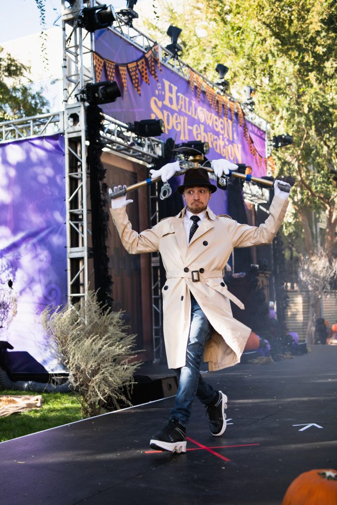 A guest dressed as Inspector Gadget walking across the Halloween Spell-ebration stage. They’re wearing a black hat with spinning fan on top and fake “arms,” as well as a long tan trench coat, blue jeans, and white shirt with black tie.