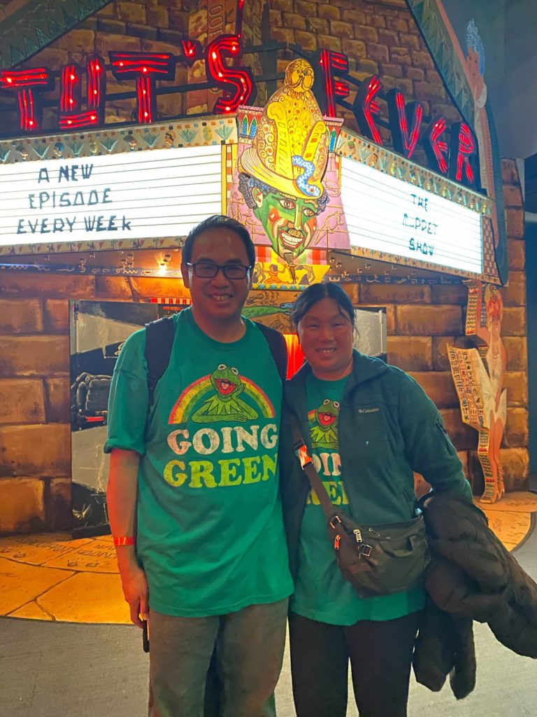 Two fans standing in front of a remake of Tuts Fever theater. The theater is a pyramid with red lettering and gold details. The fans are both wearing green shirts featuring Kermit the Frog with a rainbow behind him and reads “Going Green.”