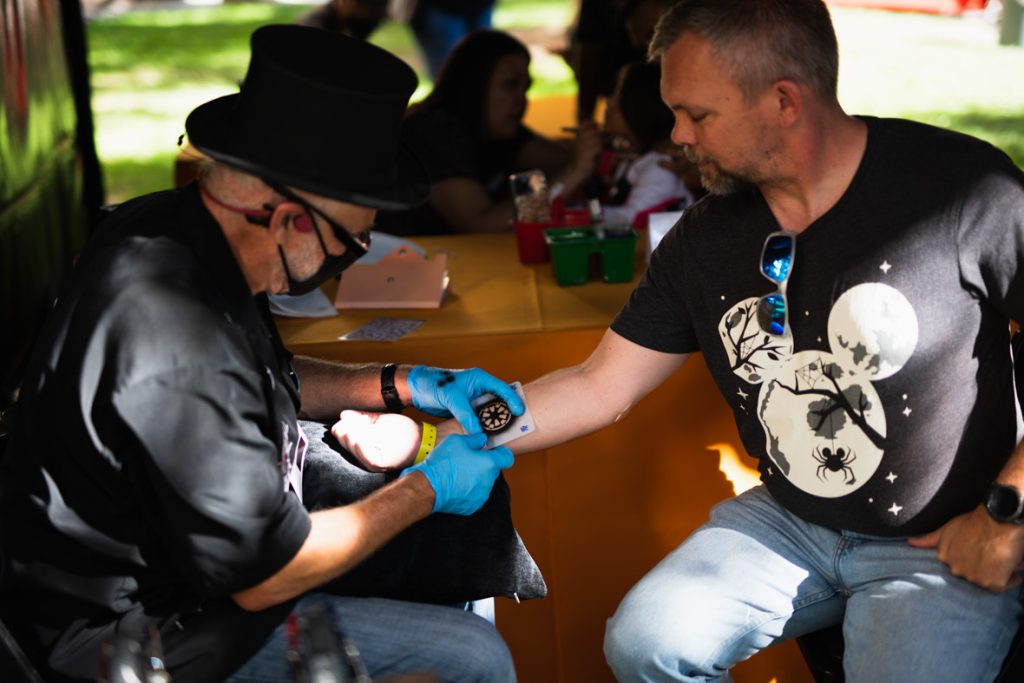 Guest getting glitter tattoo from an employee, who is wearing a black top hat, black shirt, jeans and has a grey mask and black sunglasses. The employee is putting the stencil of the tattoo on the guest’s forearm. The guest has a black t-shirt with a full moon Mickey, sunglasses, and blue jeans.