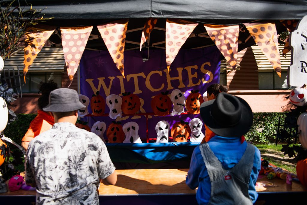 Two guests facing towards a game. The game has plush ghosts and pumpkins lined up for guests to knock down. The guest on the left is wearing a black bucket hat and a black and white shirt. The guest on the right is wearing a black cowboy hat, a blue shirt, and overalls. Above the guests’ heads are triangle banners in black and orange with polka dots.