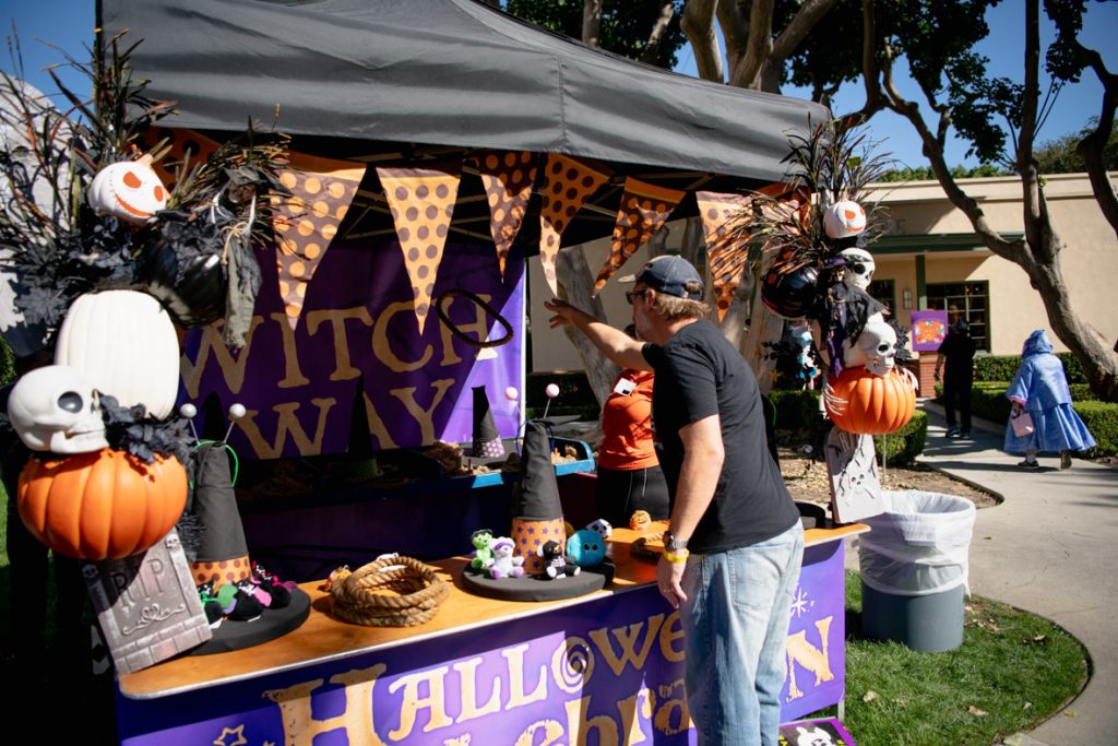 A guest throwing a ring into a game booth. They are wearing a black shirt, grey hat, and blue jeans. The booth is decorated in a variety of Halloween items including pumpkins and skulls; it also has the purple and yellow Halloween Spell-ebration logo and graphics on the front and back of the booth.