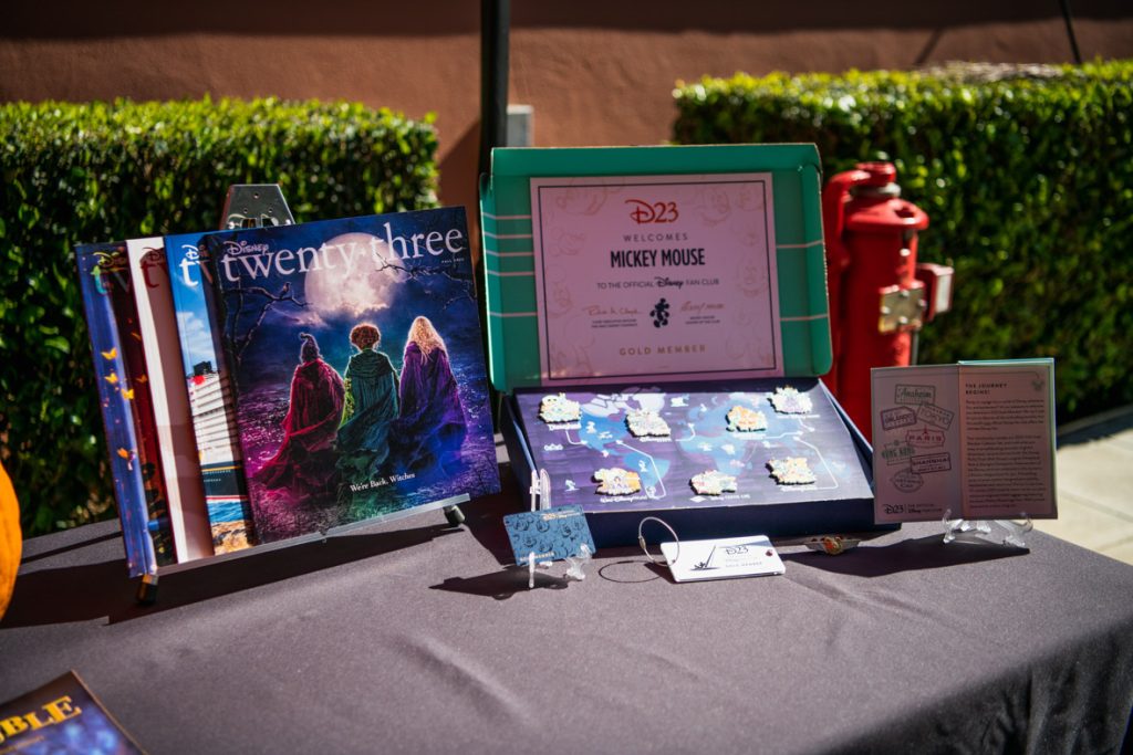 The D23 Gold Member collectors set and quarterly publications displayed on a black table. On the left, four Disney twenty-three publications are displayed overlapping each other. The front publication features the three witches from Hocus Pocus 2 facing a full moon. To the right of the publications is the pin collector set in a green and blue open box with the pins displayed. Above the pins is the D23 Gold Member certificate. In front of the pin box are the D23 Gold Member Card, silver luggage tag, gold Mickey aviator pin, and passport.