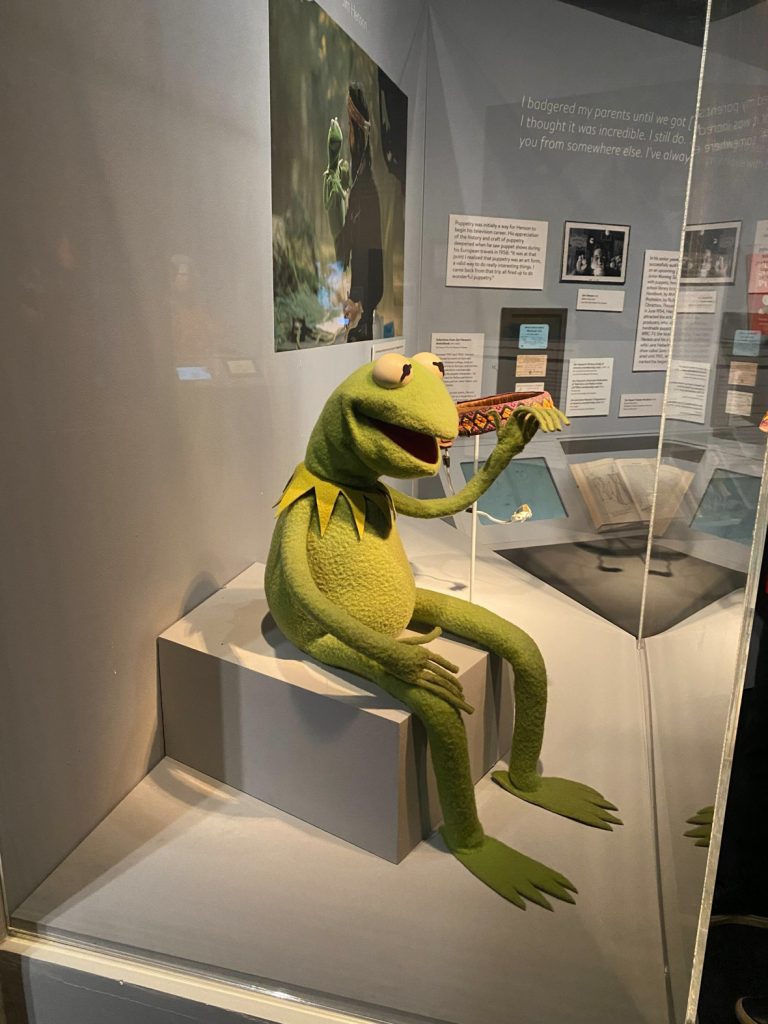A glass display box contains a Kermit the Frog puppet sitting on a white box.