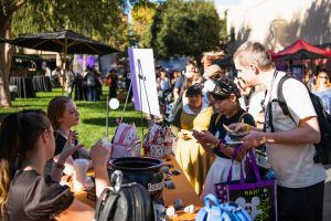 Two guests picking a prize from the Loungefly table, which has an orange tablecloth and is covered in Loungefly products and candy. Behind the table are two Loungefly workers handing out prizes to the guests. There are other guests waiting in line.