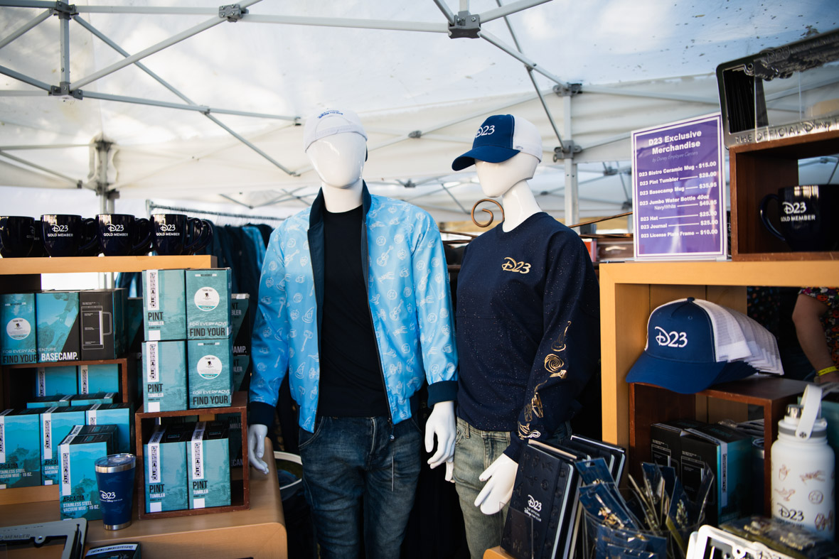 New D23 Member merchandise: On the left are black mugs with D23 Gold Member written in white letters. Below are boxes full of blue D23 tumblers with D23 franchise sketches and D23: The Official Disney Fan Club written in white. Mannequins are wearing the D23 bomber jacket in light blue with white franchise sketches on it, as well as  the D23 hat worn backwards in white with D23: The Official Disney Fan Club written in blue; the D23 Gold Member spirit jersey in dark blue with gold sparkles, gold D23 on the upper right chest, and gold sketches on the sleeve; and the D23 hat in blue with the white D23 written on the front. To the right of the mannequins are the D23 blue journals, white D23 water bottles, and D23 hats.