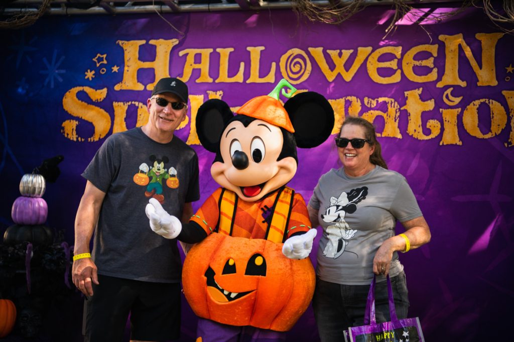 Close up photo of two guests posing with Mickey Mouse in front of a purple backdrop with “Halloween Spell-ebration” in yellow letters. The guest on the left of Mickey has a black baseball hat, grey shirt with Mickey holding two pumpkins, and black shorts. Mickey in the middle is wearing his pumpkin costume that debuted at Disneyland Resort this year. The guest to the right of Mickey has a grey shirt with Minnie dressed as Frankenstein’s bride and black leggings.