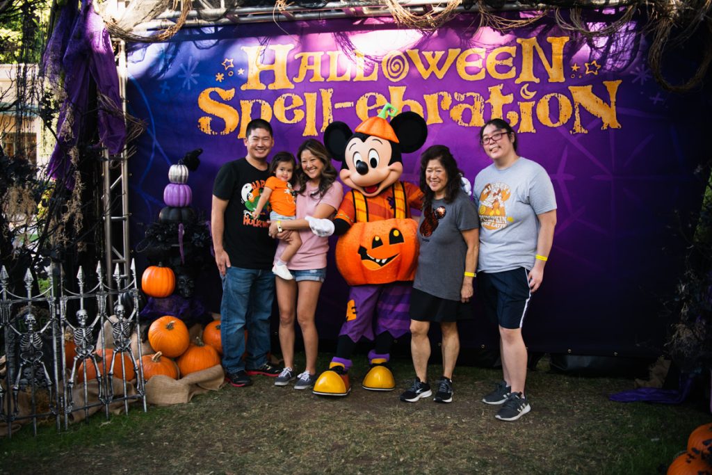 Five guests posing with Mickey Mouse in front of a purple backdrop with “Halloween Spell-ebration” in yellow letters. The guest on the right has a black shirt with a Halloween Hulk and blue jeans. The guest to the right has a pink shirt, blue shorts, and is holding a smaller guest who is wearing an orange shirt with blue shorts and white shoes. To the right of the guest is Mickey in the middle is wearing his pumpkin costume that debuted at Disneyland Resort this year. The guest to the right is wearing a grey shirt with a sequined orange Mickey pumpkin and black shorts. The guest to the right is wearing a light grey shirt with “Mickey’s Pumpkin Spice Latte” written on it and blue shorts.