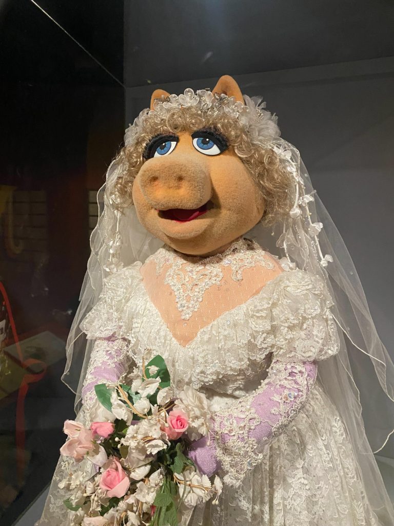 Ms. Piggy is wearing a white wedding dress with pink gloves and lace white veil and holding a bouquet of pink and white flowers.