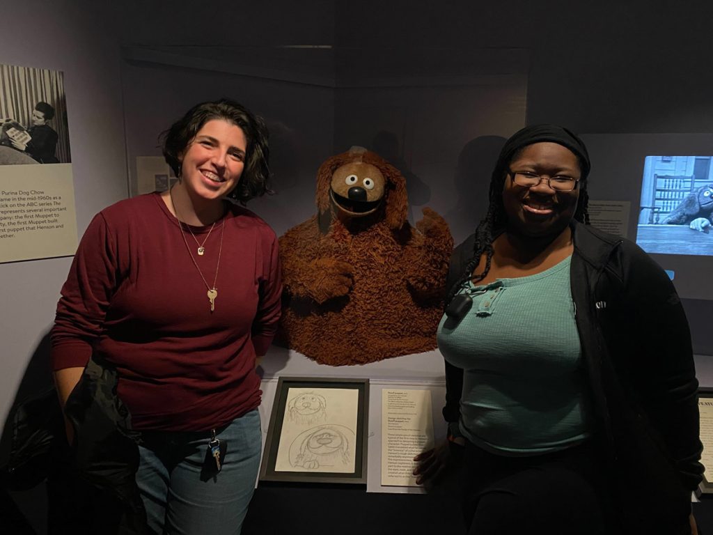 Two fans pose with Rowlf the Dog. The guest on the left of Rowlf is wearing a red sweater and blue jeans. The guest on the right of Rowlf is wearing a blue shirt, black jacket, and black jeans.
