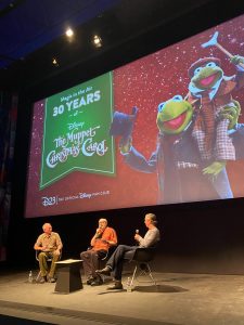 Three speakers sit on a stage in front of the event logo. The event logo features a deep red background covered with swirls and snow. On the left is a green banner reading, “Magic in the Air. 30 Years of The Muppet Christmas Carol.” Beneath the green banner is a white D23: The Official Disney Fan Club logo. To the right of the image is Kermit with Robin the Frog on his shoulder. Kermit is dressed in his Christmas Carol attire with a black top hat held in his right hand, a long gray coat, checkered shirt, and red and black striped scarf. Robin the Frog is dressed as the character Tiny Tim with a red hat, orange coat, white shirt, plaid orange pants, and orange jacket. He is holding his brown cane in his right hand. The speakers below the projected logo are (from left to right) Fred Buchholz, Craig Shemin, and Peter MacKennan. Frank is wearing a tan button up shirt and green pants and is holding a microphone. Craig is wearing an orange button up shirt with brown pants and is speaking into a microphone. Peter is wearing a long grey shirt with grey pants and is holding a microphone.