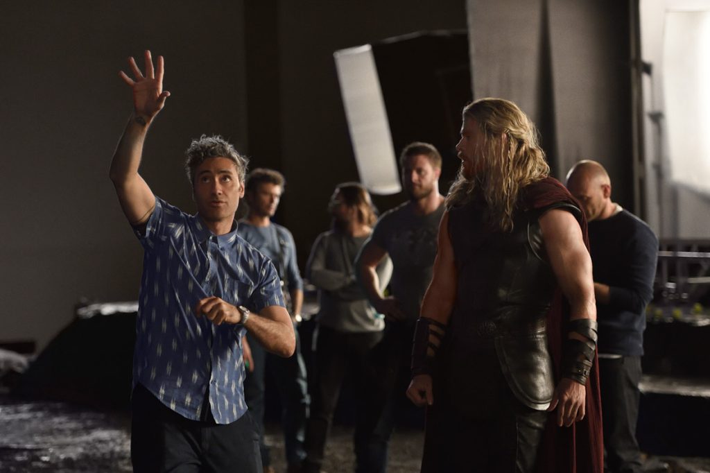 Thor: Ragnarok director Taika Waititi raises his hand up, illustrating something to actor Chris Hemsworth as they stand together on set.