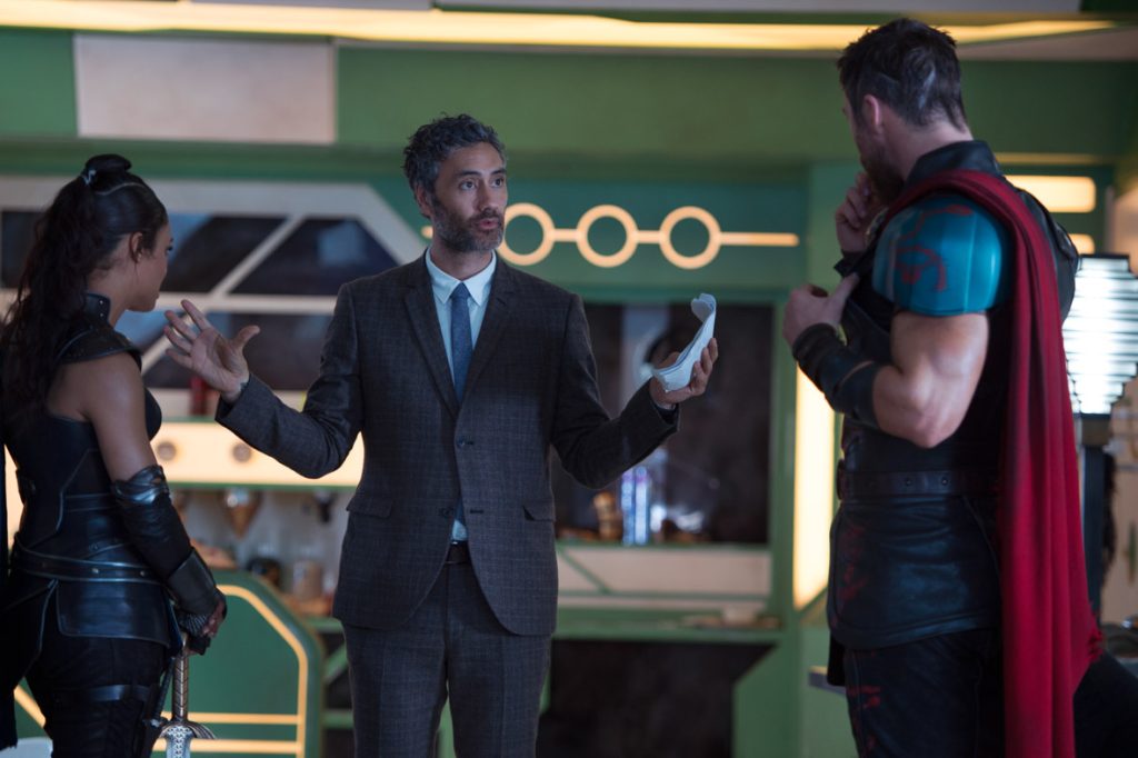 Thor: Ragnarok director Taika Waititi stands between actors Tessa Thompson and Chris Hemsworth on set. Both actors have their back turned to the camera, facing Waititi as he directs them.