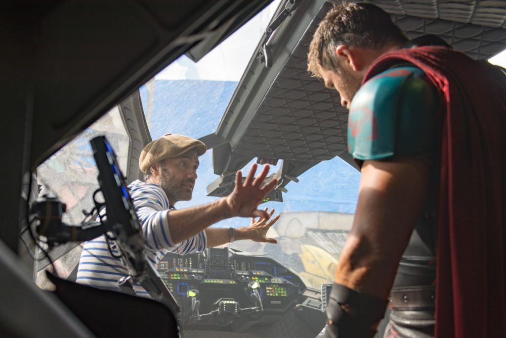 Chris Hemsworth stands inside the Avengers’ Quinjet, his back to the camera. Facing Hemsworth, Taika Waititi directs the actor with outspread arms.