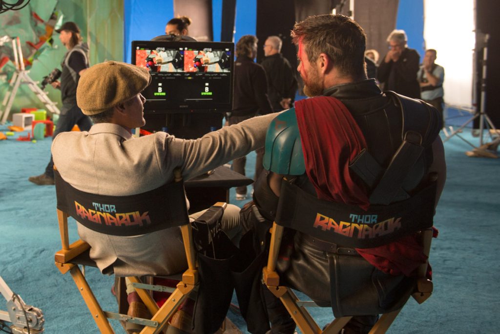 Thor: Ragnarok director Taika Waititi and actor Chris Hemsworth sit in Thor: Ragnarok-labelled directors chairs, watching footage from the day on a small monitor. Their backs are to the camera, and Waititi is pointing across Hemsworth at something off screen.