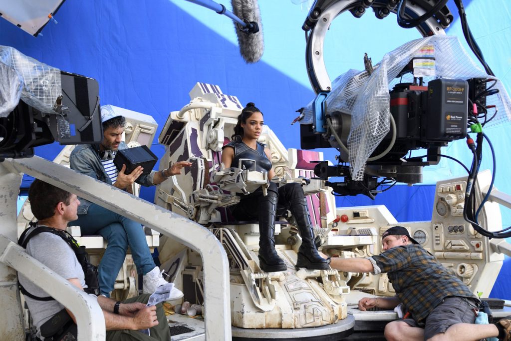 Actress Tessa Thompson sits in a set backed by a blue screen, representing the cockpit of her character’s ship. A camera faces her, as various crew members prepare her and the set for the next shot. Director Taika Waititi sits next to her, legs crossed, as he watches her through a handheld camera.