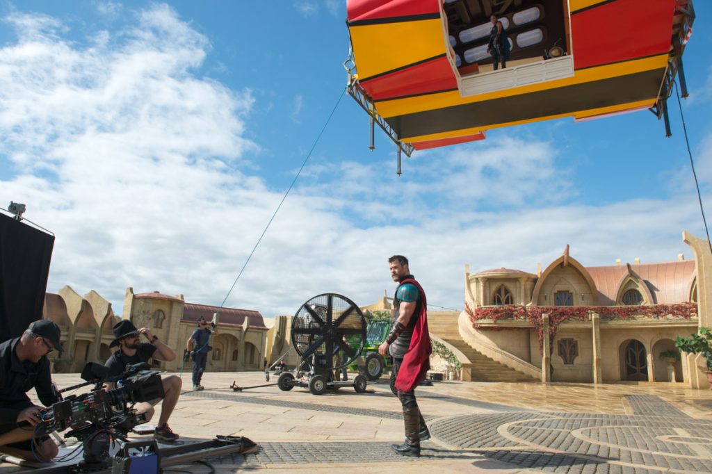 Chris Hemsworth stands in the center of the Thor: Ragnarok Asgard set, while Tessa Thompson stands above him in an elevated room that will be transformed into a spaceship via visual effects. Film crew stand off to the left of the image, preparing for the next shot.