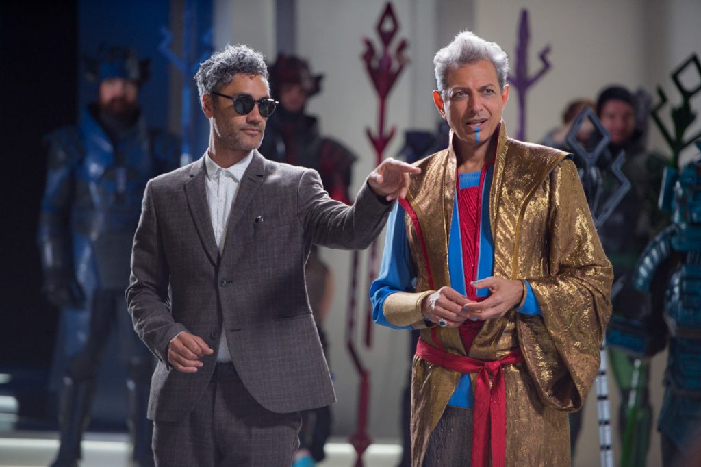 Thor: Ragnarok director Taika Waititi, in a suit and sunglasses, walks along with actor Jeff Goldblum, in full costume as The Grandmaster. Waititi is pointing at something off to the right.