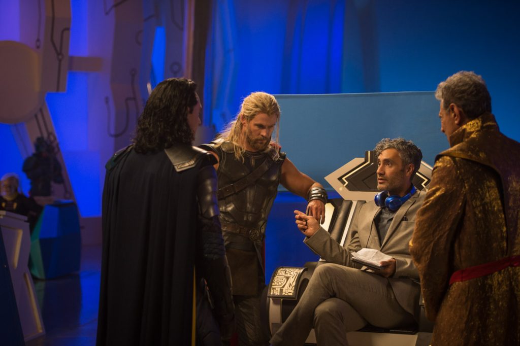 Thor: Ragnarok director Taika Waititi sits on The Grandmaster’s throne, headphones around his neck, as he talks with actors Tom Hiddleston, Chris Hemsworth, and Jeff Goldblum. Hiddleston and Goldblum both have their backs to the camera, while Hemsworth faces the viewers, arm resting on the side of the throne.