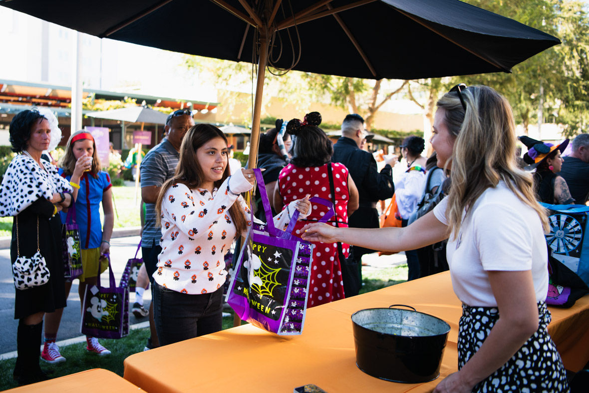 A guest holding an open bag for an employee to fill with candy. The guest has long dark hair and is wearing a white shirt with Jack Skellington heads. The employee handing the candy across the table has long blonde hair and is wearing a white shirt and polka dot skirt. The trick-or-treat bag is purple with a Mickey Mouse ghost and “Happy Halloween” written in green.