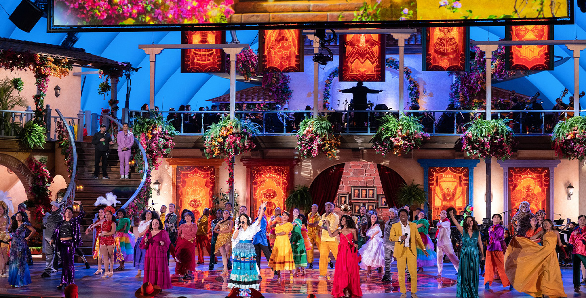 A promo image for Encanto at the Hollywood Bowl, airing on Disney+ December 28. The entire cast is seen singing and dancing in colorful outfits, spread out on the stage, which is set up like the two-story Casa Madrigal from the film; on the second “story” sits the orchestra, seen mostly in silhouette. There is a stairway seen to the left, leading to the second “story.” Hanging plants line where the first and second “stories” divide.