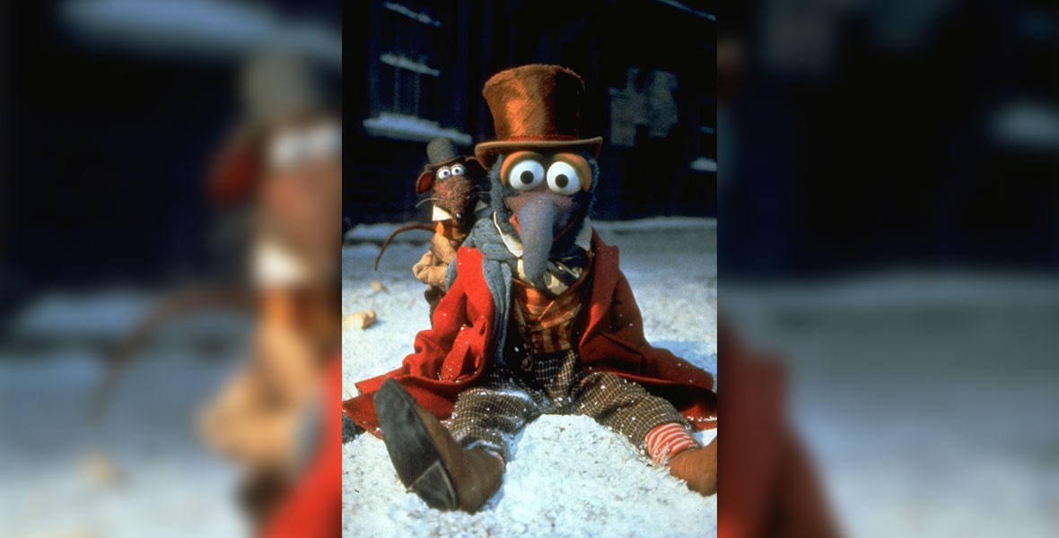 In an image from The Muppet Christmas Carol, Gonzo as Charles Dickens is wearing an orange-ish top hat, plaid pants, and a red overcoat, and is sitting in the snow, with Rizzo the Rat (wearing a black top hat) looking over his left shoulder.