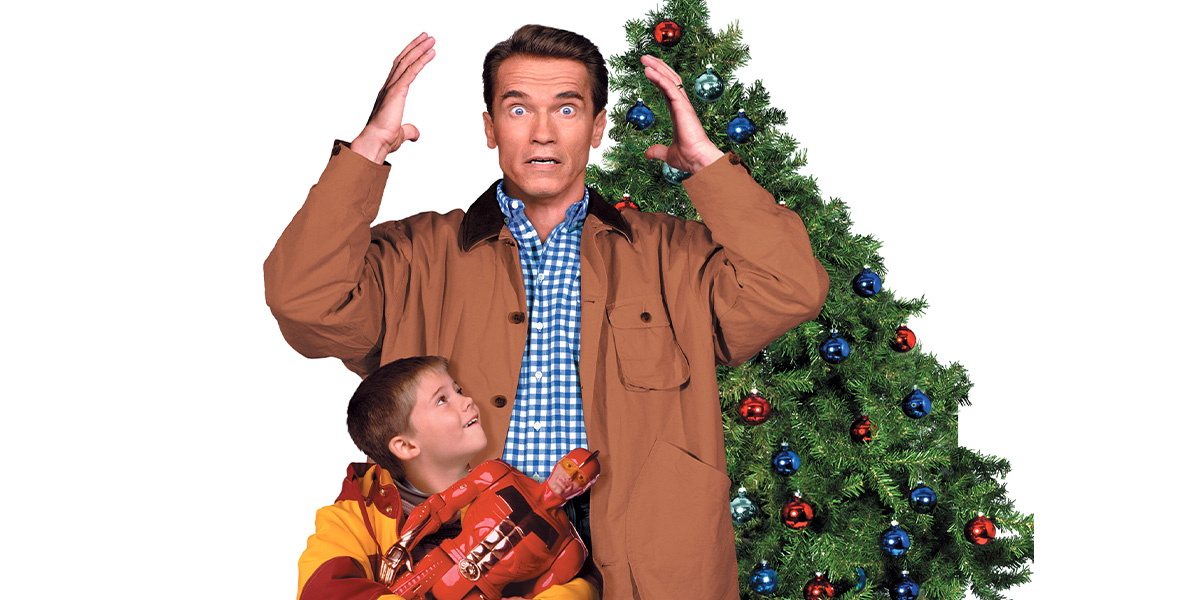 In the key art for the 20th Century Studios film Jingle All the Way, actor Arnold Schwarzenegger portrays Howard Langston and stands wide-eyed with his hands on each side of his head. He wears a blue gingham button-down shirt, tan coat, and dark blue denim pants. Actor Jake Lloyd portrays Jamie Langston and looks up at Schwarzenegger while holding a large male action figure; he wears a red and yellow striped jacket. They’re both standing in front of a green Christmas tree covered with shiny bright red, light green, and royal blue circular Christmas ornaments.