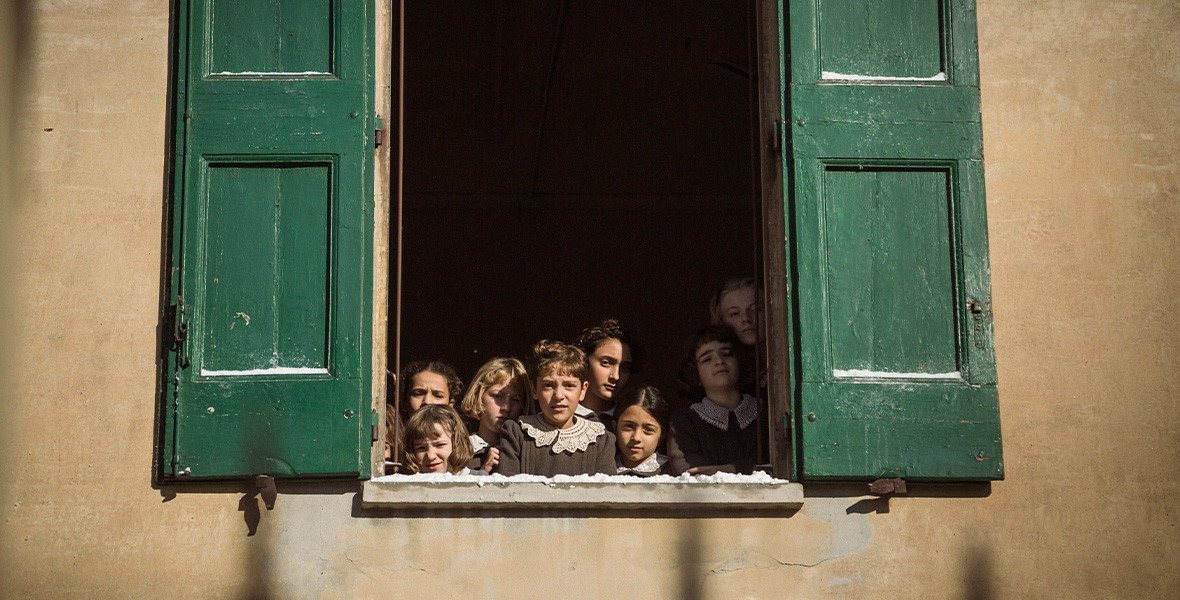 In a scene from Le Pupille, a group of young girls stand huddled together and look outside a large window. The girls wear simple brown dresses adorned with cream lace collars. On each side of the window hang large, hunter green, wooden shutters.
