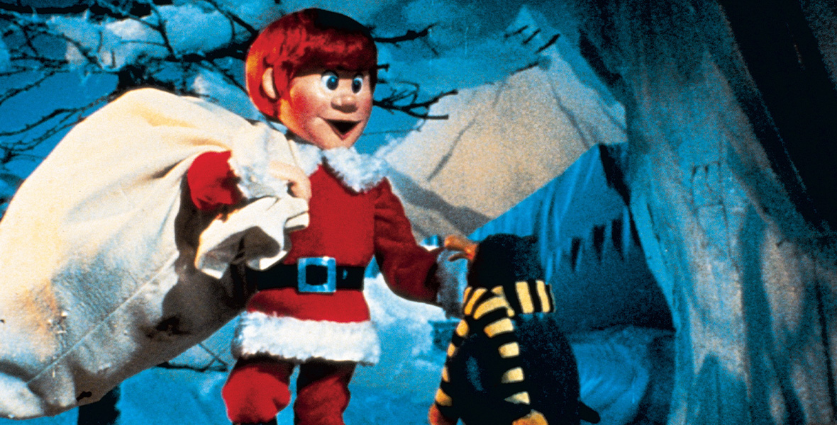 In a scene from Santa Claus Is Comin’ to Town, a stop-motion red-haired male character wears a Santa Claus suit and holds a large canvas sack over his shoulder. He faces a penguin that wears a black and yellow scarf around its neck. Both are surrounded by snow and icicle covered trees.