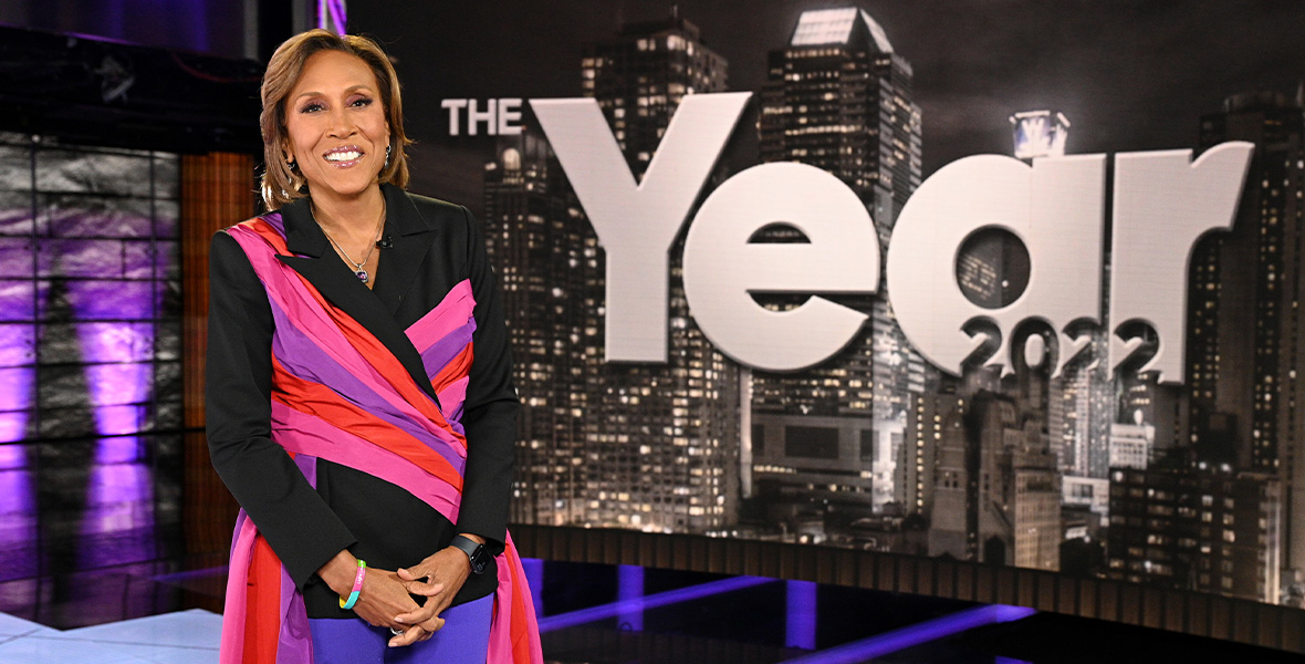 In a scene from the ABC News special The Year: 2022, host and Disney Legend Robin Roberts stands with her arms folded in front of her and in front of a large television screen with the show’s title. Roberts wears a black blazer jacket with pink, red, and purple fabric crisscrossing and cascading down her sides, and a pair of purple pants.