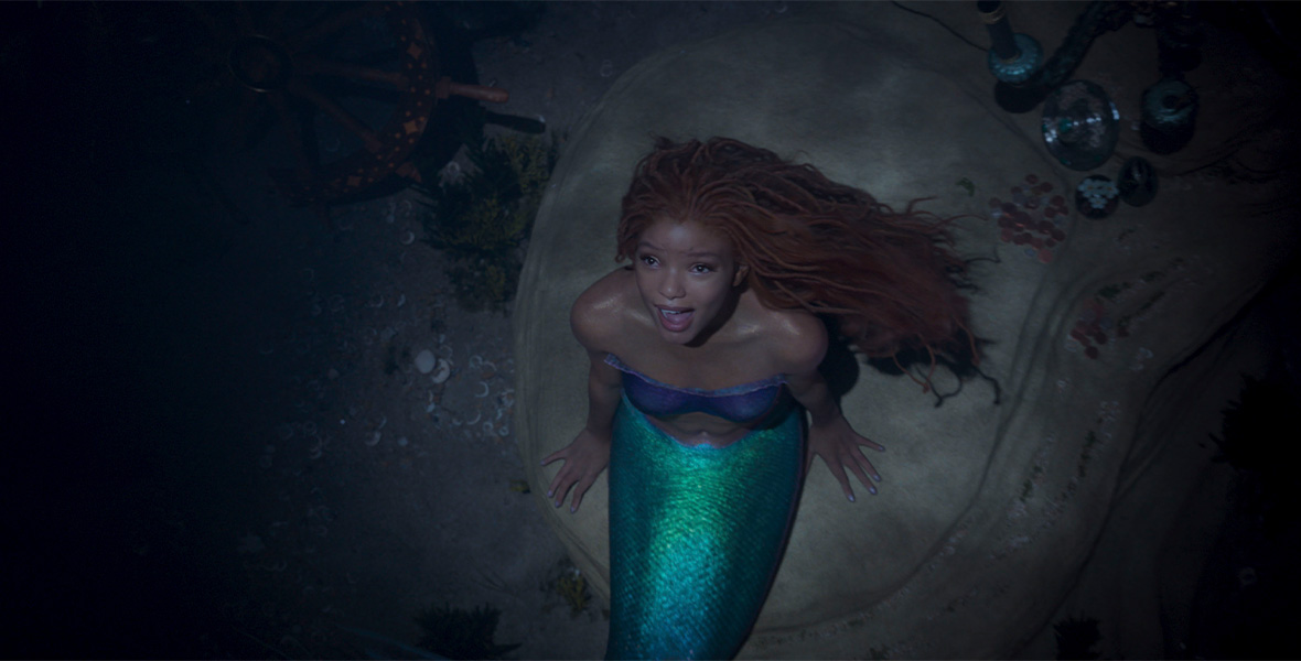In a scene from Walt Disney Studios’ The Little Mermaid, actor Halle Bailey portrays Ariel, a mermaid with long, flowy red hair, an iridescent green mermaid tale, and an iridescent purple strapless top. Bailey looks up and sits on a large rock deep below the ocean’s surface.