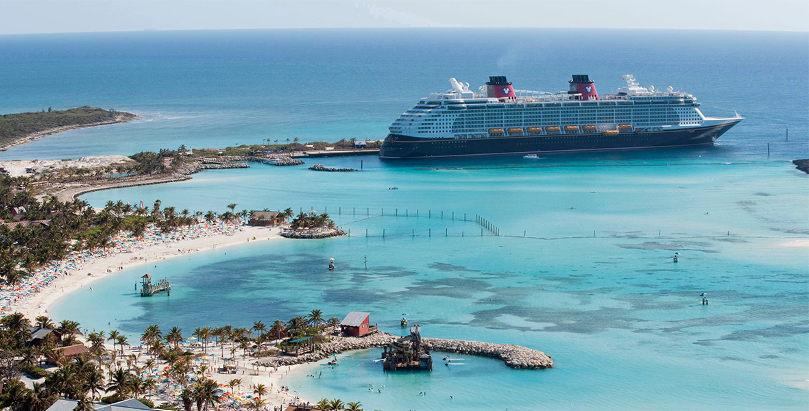 Image of Disney’s island, Castaway Cay, from above. A Disney Cruise Line ship is seen at the top right of the image; on the left and bottom are several inlets, where there are beaches filled with people and umbrellas. Further inland are many trees, huts, and other buildings. The water is a bright blue.