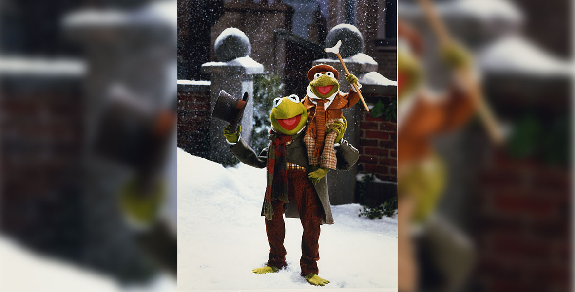 In an image from The Muppet Christmas Carol, Kermit the Frog as Bob Cratchit is standing in the snow while wearing a gray overcoat and red plaid pants, and holding a gray top hat in one hand. He’s also holding Robin as Tiny Tim—who’s wearing an orange hat and orange plaid pants and a coat—up on his right shoulder. Tiny Tim is holding a crutch in his right hand.