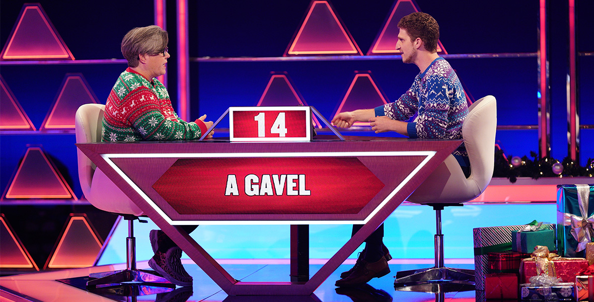In a scene from ABC series The $100,000 Pyramid, actor and comedian Rosie O’Donnell sits at a table across from a contestant. She wears black framed glasses and a green and red striped Christmas sweater with white snowflakes, polka dots, and reindeers. The constant wears a blue and purple Christmas sweater with white snowflakes, chevrons, and reindeers. The desk is shaped like an upside-down pyramid and has two digital screens. The top, smaller screen has a red background and reads “14” in white. The larger screen, also with a red background, reads “A GAVEL” in white. Behind O’Donnell and the contestant are pink and purple led lights in triangle shapes.