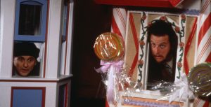 In a scene from 20th Century Studios&rsquo; Home Alone 2: Lost in New York, actors Joe Pesci and Daniel Stern each look out of the windows of two miniature houses with Christmas decorations. Pesci wears a solid black beanie and black coat. Pesci stands inside a small, white house with blue windows and red trim. Stern wears a white T-shirt and black jacket. Stern stands inside a miniature house decorated like a gingerbread house with a colorful roof, candy cane-like pillars, red trim, and circular, colorful lollipops.