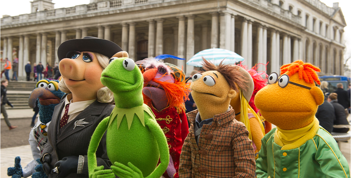 In a scene from Muppets Most Wanted, Muppets Gonzo, Miss Piggy, Kermit the Frog, Floyd Pepper, Walter, and Scooter stand in a cluster and look to the left. Behind the group is a large, ornate, stone building. Miss Piggy wears a gray fedora, a gray suit with white pinstripes, a tan blouse, and a cardinal tie with white polka dots. Walter wears a brown, green, and orange plaid button-down shirt. Scooter wears a yellow turtleneck and a shiny green bomber jacket.