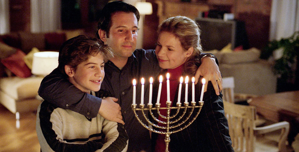 In a scene from Disney’s Full-Court Miracle, actor Jason Blicker as Marshall Schlotsky wraps his arms around actors Alex D. Linz as Alex Schlotsky and Linda Kash as Cynthia Schlotsky. The three characters look at a lit menorah with white candles on a white countertop. Blicker and Linz both wear black yarmulkes atop their heads. Linz wears a tan sweater with brown stripes down the sleeves. Blicker wears a black button-down shirt. Kash wears a red turtleneck sweater and a black jacket.