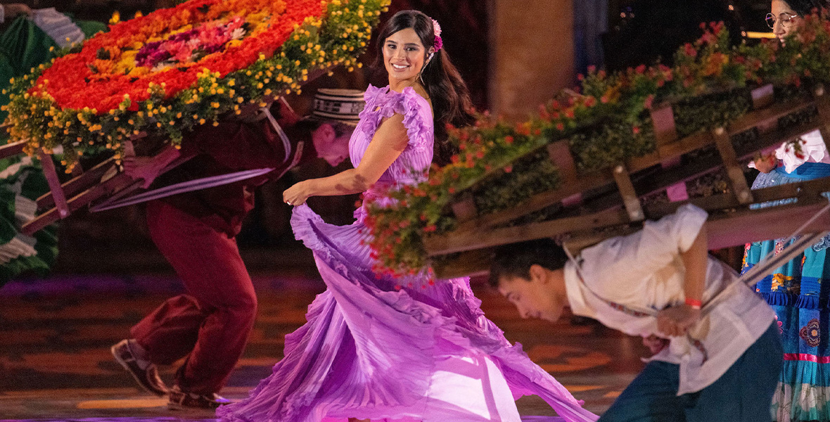 In a scene from Encanto at the Hollywood Bowl, actor Diane Guerrero portrays Isabella and dances on stage surrounded by platforms of colorful flowers. Guerrero wears a bright purple flowy dress and a pair of iridescent, purple platform heels.