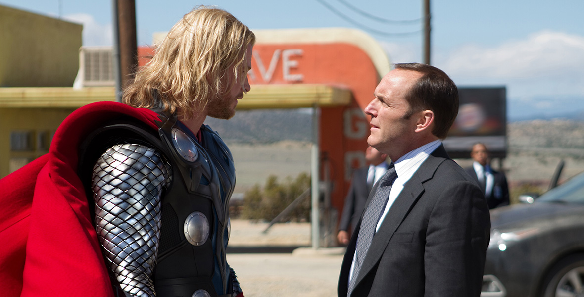 In a scene from Marvel Studios’ Thor, actor Chris Hemsworth portrays Thor and actor Clark Gregg portrays Agent Coulson. Hemsworth and Gregg stand face-to-face in a desert landscape next to a vintage electronic sign painted yellow and orange. Hemsworth wears silver and black armor and a red cape; Gregg wears a black suit, a black tie, and a white button-down shirt.