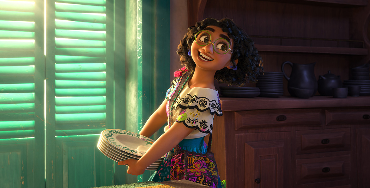 Encanto’s Mirabel is holding a stack of plates while looking over her left shoulder. She is wearing a white layered shirt and glasses.