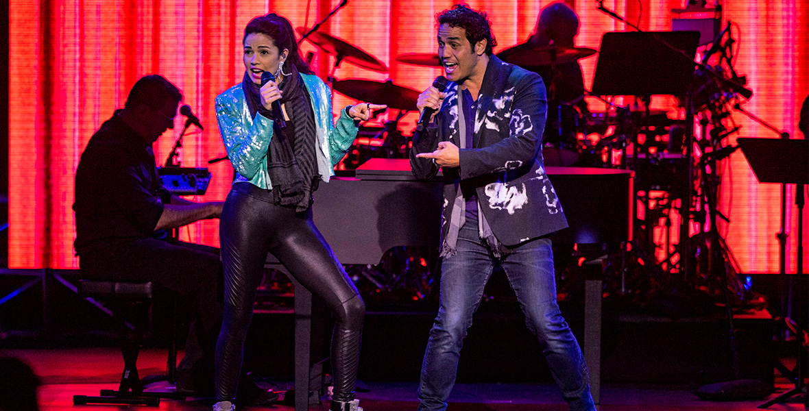 Broadway performers Arielle Jacobs and Adam Jacobs on stage, mid-song, in a promotional image for the 2023 DISNEY ON BROADWAY concert series at EPCOT, during the International Festival of the Arts. Arielle (on left) is wearing a shiny turquoise jacket, black leggings, and sparkly silver hi-top sneakers; Adam (on right) is wearing a black and white patterned suit jacket and grey scarf with dark jeans. A piano player and drummer are silhouetted behind them, with an orange and red lit-up background.
