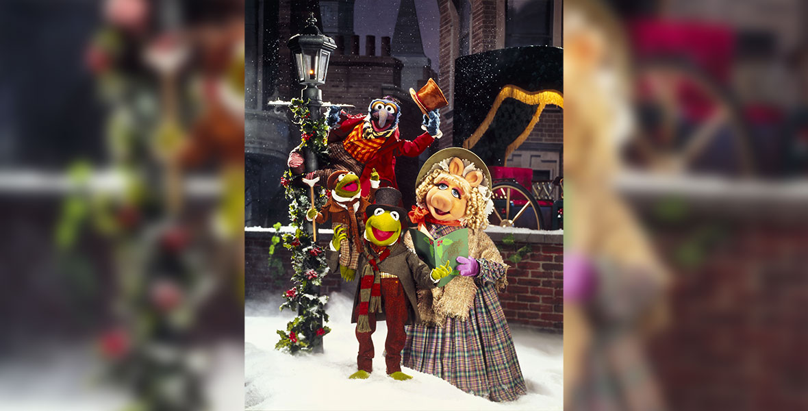 In an image from The Muppet Christmas Carol, from upper left to right, Gonzo as Charles Dickens, Robin as Tiny Tim, Kermit the Frog as Bob Cratchit, and Miss Piggy as Emily Cratchit are standing in the snow near a holly-wrapped streetlamp; Gonzo is holding onto the lamp itself, and Robin is sitting on Kermit’s left shoulder. Miss Piggy is holding a book of Christmas carols. They are all dressed in Victorian holiday finery, including top hats, a bonnet for Miss Piggy, and scarves.