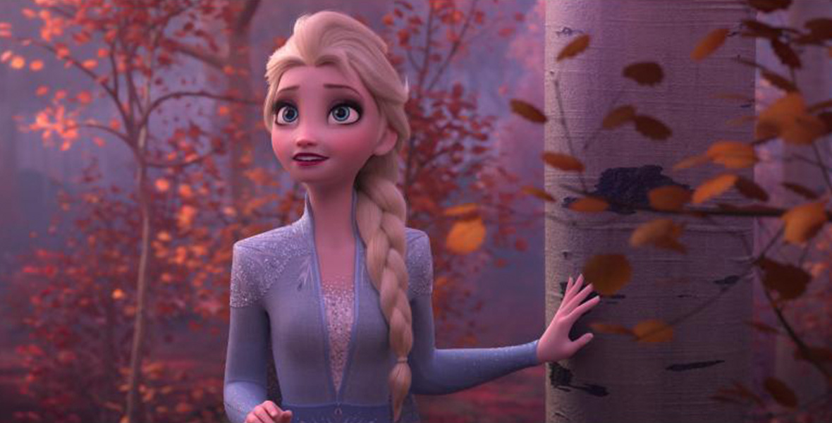In a scene from Walt Disney Animation’s feature film Frozen 2, Elsa stands in forest with several brown and orange trees, with her left hand placed on a light brown tree. Elsa has long blonde hair that is styled in a large single braid, big blue eyes, and wears a light blue, long-sleeved dress with white embroidery on each shoulder.