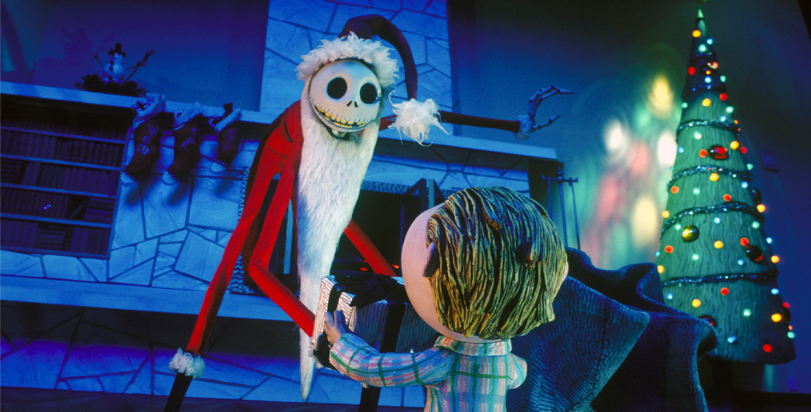 In a scene from Tim Burton’s The Nightmare Before Christmas, Jack Skellington, a stop-motion skeleton man, wears a red Santa Claus suit, a long, white beard, and a pair of tall black boots. Skellington hunches forward to speak to a young boy. Both stand in a dimly lit room that is decorated with a green Christmas tree covered in colorful lights.