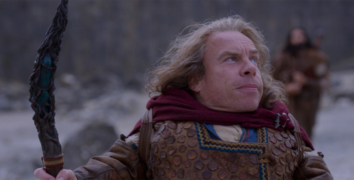 In a scene from Disney+ Original series Willow, actor Warwick Davis portrays Willow Ufgood. Davis holds a carved wooden staff in his right hand and wears a maroon cape, a brown textured coat, and a tan long-sleeved shirt. Behind Davis is a snow-covered forest.